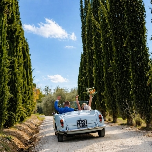 Bruidspaar rijdt in een cabrio tussen de cipressen in Toscane.